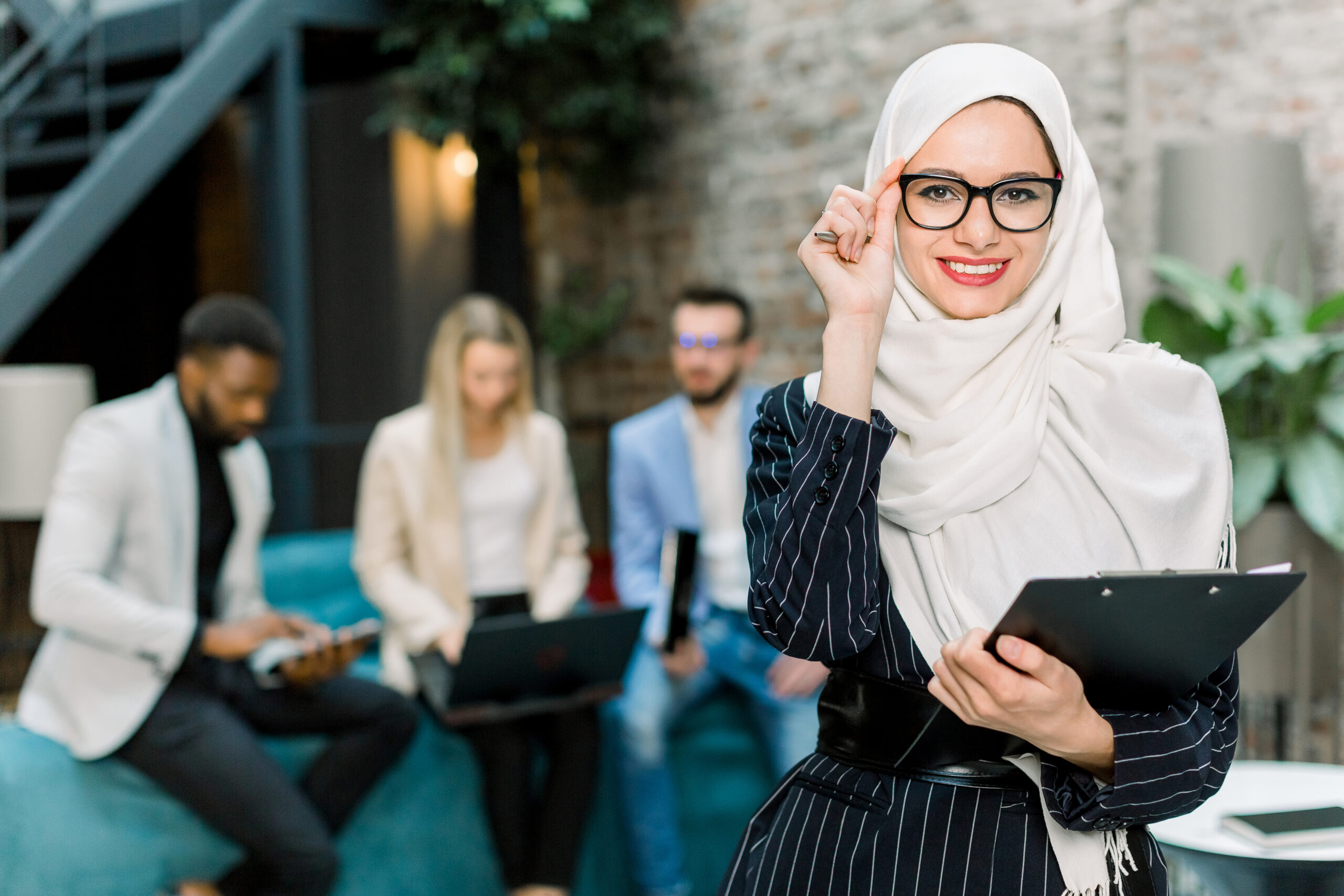 Young pretty Muslim business woman in white hijab and eyeglasses, standing in modern office, holding folder with papers and smiling to camera. Three multiethnic office workers on the background